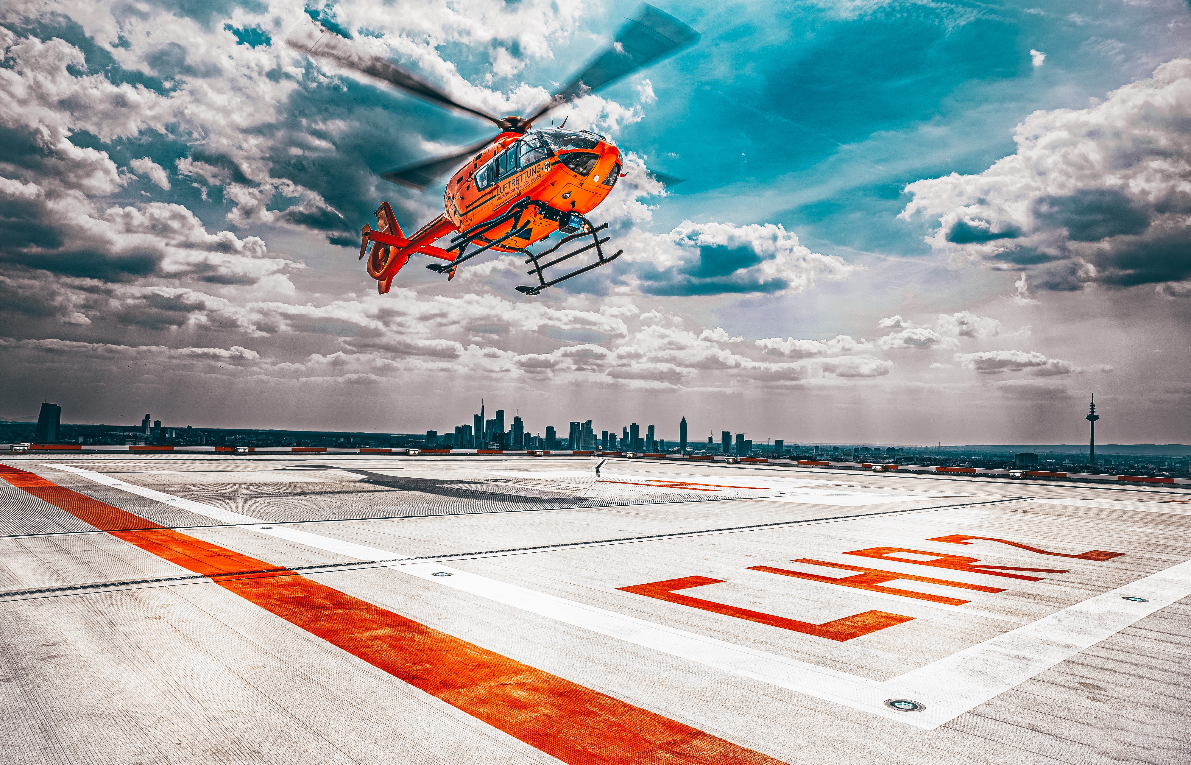Blick auf ein Hausdach auf dem sich ein Landeplatz befindet. Ein orangefarbener Hubschrauber setzt zur Landung an. Der Himmel ist blau und voller Wolken. Im Hintergrund ist die Skyline von Frankfurt am Main zu erkennen.