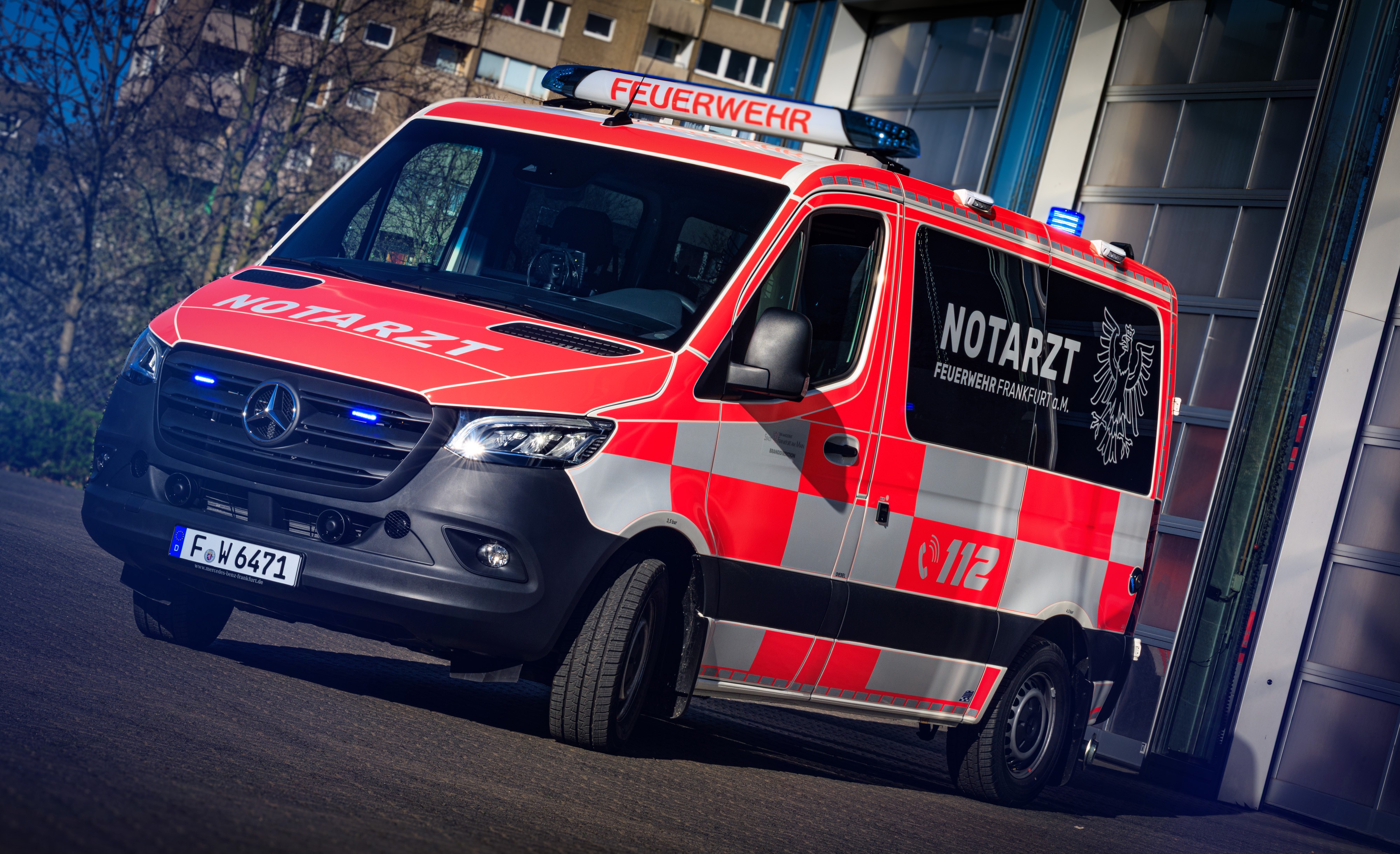Ein orangefarbenes Notarztfahrzeug steht auf der Straße. Auf dem Dach des Wagens befinden sich Blaulichter und ein Schild mit der Aufschrift Feuerwehr. Auf der dunkel gefärbten Scheibe ist die Aufschrift Notarzt und Feuer Frankfurt a. M. Angebracht.