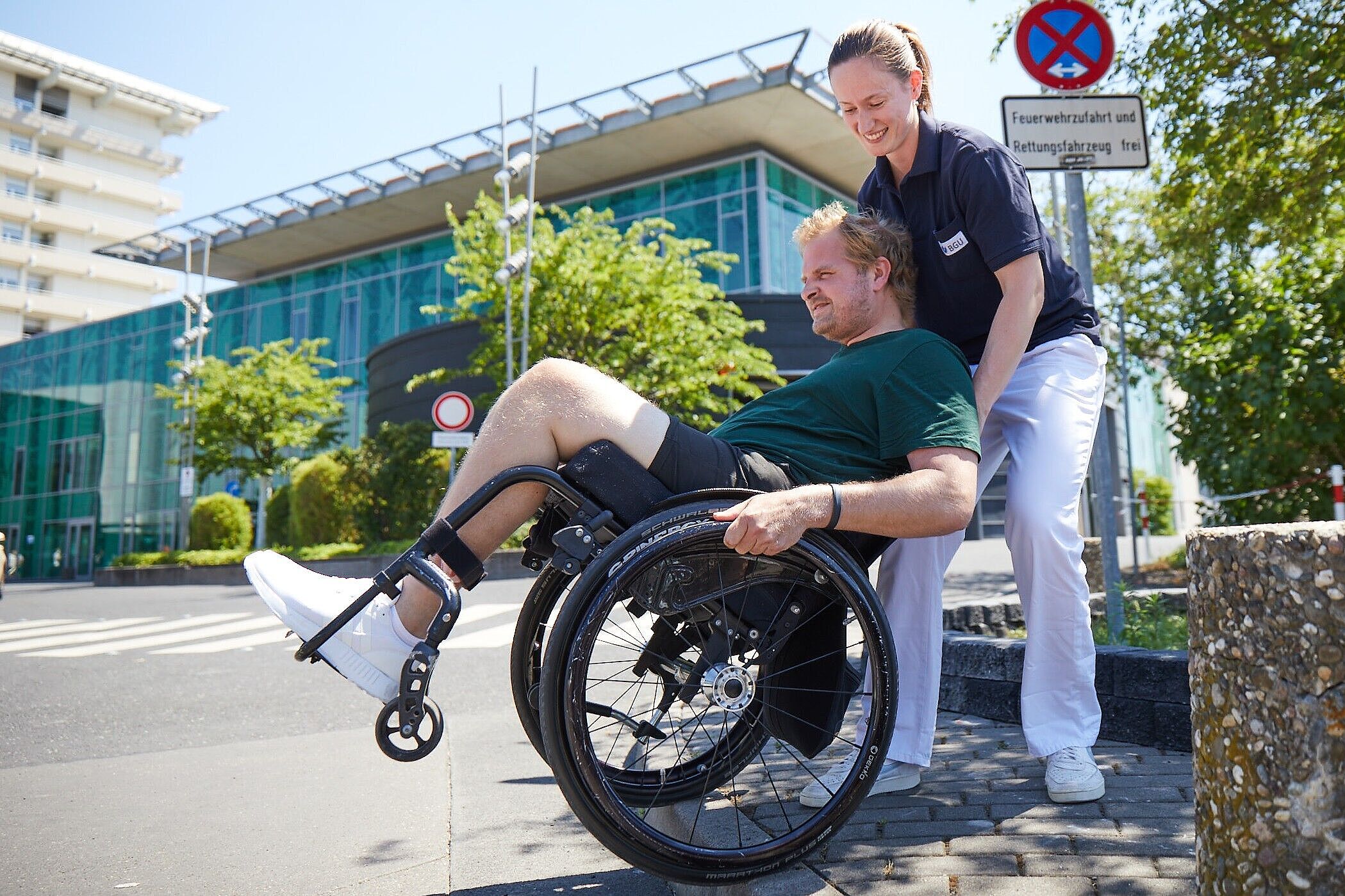 Therapeutin mit einem Patienten im Rollstuhl vor Klinikgebäude